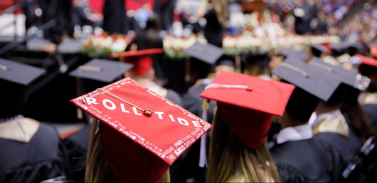 A red honors graduation cap is seen decorated with a white "Roll Tide" at a commencement ceremony.