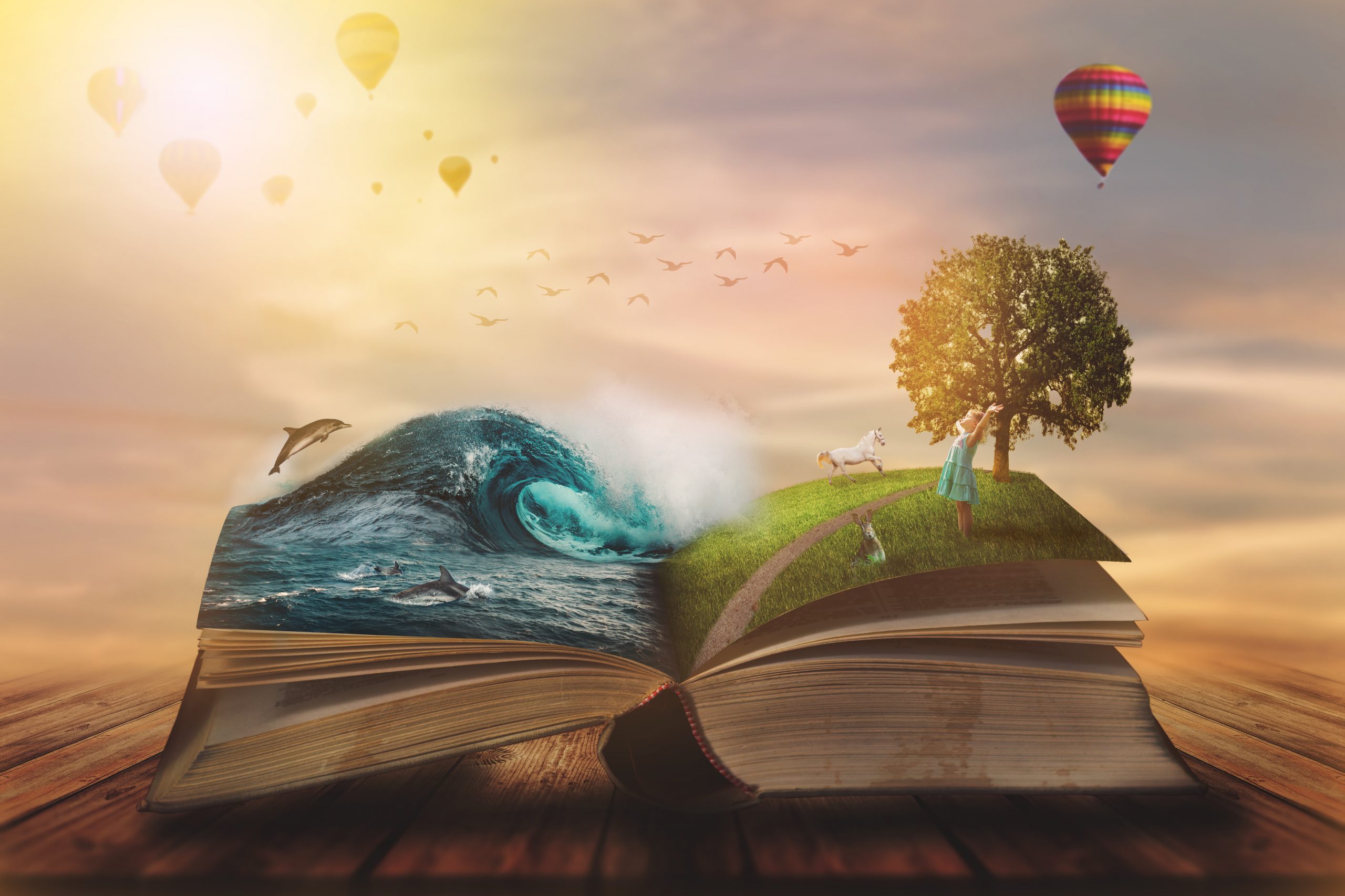 An open book rests on a wooden surface with its pages transforming into a fantastical scene. On the left, a large ocean wave rises from the pages with dolphins leaping from the water. On the right, a grassy hill with a tree, a girl in a dress, and a white horse emerges from the book. Hot air balloons float in a glowing sky, and birds fly across the horizon, creating a dreamy, surreal atmosphere.