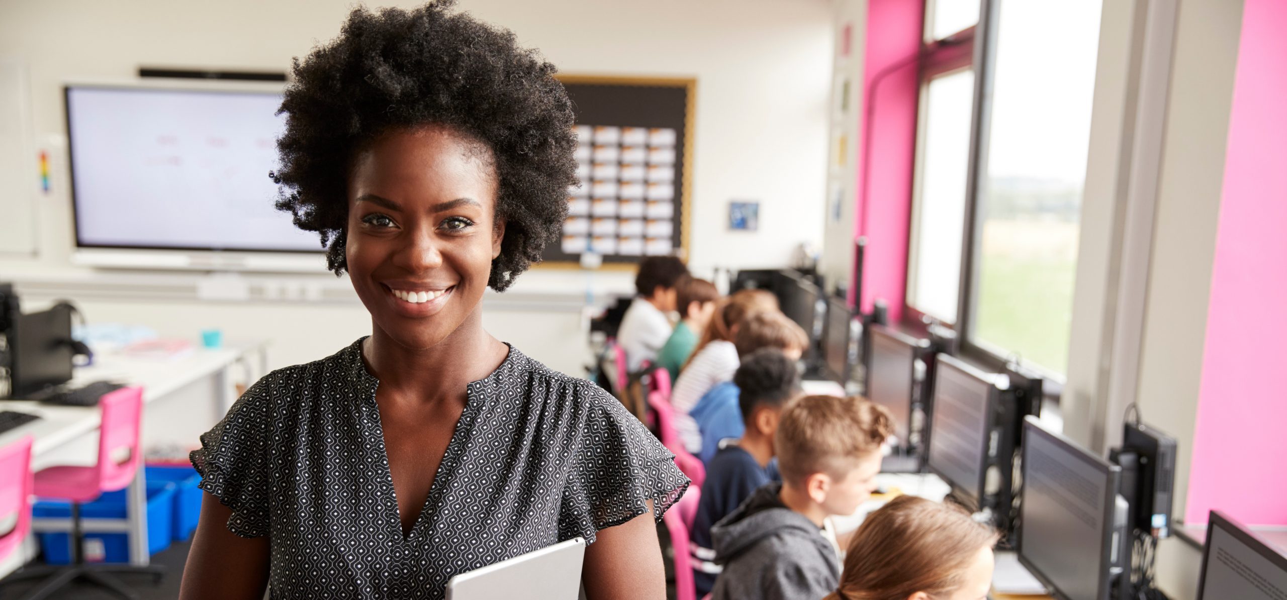 Teacher holds a tablet as students are working on computers behind her.