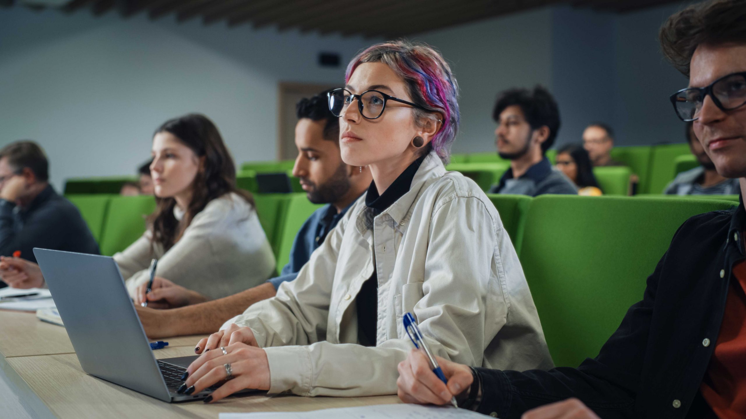 A young woman with colorful hair sits in an auditorium classroom, taking notes on her laptop.